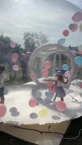 A clear, inflatable dome or bubble is filled with many colorful balloons. Inside, two children, a boy and a girl, are playing. The boy, wearing a striped t-shirt and shorts, stands on the left side of the dome. The girl, wearing a white top and a light purple skirt and hat, is in the center, running towards the right. Several people are visible outside the dome, standing in a grassy, outdoor area. The sky is bright and cloudy, suggesting daytime. The dome has a white, inflated floor and an inner inflatable structure. Balloons of various colors, including pink, yellow, blue, and green, are scattered both inside and outside the dome. The overall scene depicts a playful outdoor event.