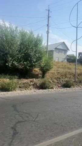The video captures a roadside view of a residential area under a clear sky. The camera pans across a slightly elevated landscape with sparse vegetation, featuring several houses in various stages of construction or completion. A partially built brick structure is visible, along with a finished house featuring a light-colored facade and a darker roof. Another larger house with a modern design and large windows is also present. In the foreground, a paved road with visible cracks runs horizontally across the frame. To the right, a metallic pole with curved elements stands near the road. Further in the background, a cluster of tall trees creates a green backdrop, and beyond that, what appears to be a fenced sports field or recreational area with small buildings and structures is discernible. A green tractor is parked on the incline near one of the houses. The overall impression is of a developing rural or semi-urban environment.