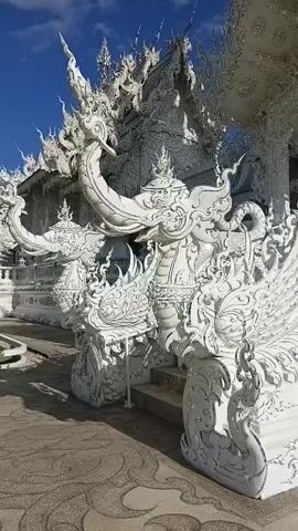 A video displays intricate white architectural carvings and sculptures of mythical creatures, possibly dragons and birds, against a bright blue sky. The scene appears to be an outdoor location, with detailed stonework on a staircase and facade. The camera pans across the elaborate, ornate designs, highlighting the texture and detail of the white structures.
