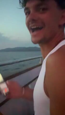 The video shows a young man with dark hair and a white tank top, smiling broadly and looking at the camera. He is holding a smartphone in his left hand. In the background, there is a body of water and a blurry landscape of hills or mountains. The sky appears to be dusk or dawn. The video then shifts to show a different scene on a boat. People are visible on the upper deck of the boat, and there is a set of stairs leading up to it. The lighting suggests it is evening.