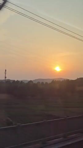 The video shows a sunset over a rural landscape. The sun is a bright orange orb low in the sky. Silhouetted against the sunset are rolling hills covered in trees. In the foreground, there are power lines stretching across the frame. A tall metal power line tower is visible in the distance, as is a smaller telecommunications tower. Fields of dark green and brown vegetation are visible between the power line tower and the hills. The video appears to be filmed from a moving vehicle, as indicated by the slight blur and the way the power lines move across the frame.