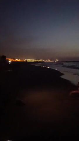 This media file captures a group of people enjoying the sea at Port-la-Nouvelle, France, during twilight. The scene unfolds on a dark beach as a dusky sky transitions from deep blue to shades of orange and pink near the horizon. The sea is calm with gentle waves lapping at the shore, and the lights of the distant port shimmer across the water.

The primary subjects are several individuals, appearing as silhouettes against the dimly lit water, splashing and playing in the shallow sea. Their forms are indistinct due to the low light, but their energetic movements suggest a sense of fun and freedom. The overall atmosphere is one of peaceful evening enjoyment, with the fading light creating a serene and slightly magical ambiance. The location cues, such as the distant lights of a port, are subtle but confirm the setting as a coastal area with human activity nearby. The time of day is clearly dusk or early evening, shortly after sunset. There is no visible text in the media file.