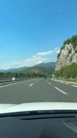 This is a first-person perspective video recorded from inside a car driving on a highway. The road is paved with asphalt and has white lane markings. To the left, there is a guardrail separating the highway from a vegetated area, and in the distance, mountains are visible. To the right, a rocky cliff face is prominent, with some greenery growing on it. The sky is a clear, bright blue with a few scattered white clouds, suggesting a sunny day.

Several road signs are visible. One sign indicates "km 64". Another green sign displays "E 65". There are also gantries or overhead structures visible along the highway, which may be associated with traffic management or electronic toll collection. The overall scene depicts a scenic drive through a mountainous and somewhat rural landscape. The time of day appears to be daytime, likely late morning or early afternoon, given the bright sunlight. The weather seems pleasant and clear, ideal for travel. The atmosphere conveyed is one of movement and a journey through natural surroundings.