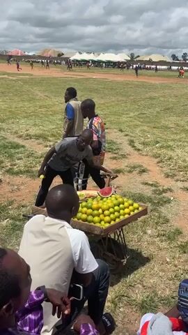 The video shows an outdoor event, possibly a sports match, with a field in the background where people are playing. There are tents and structures in the distance. In the foreground, there are several people. One man is standing next to a cart filled with oranges and a watermelon. He is holding a yellow object, possibly a lemon. Two other men are standing nearby. One is wearing a gray shirt and black pants, and the other is wearing a colorful striped shirt and black pants. Further in the foreground, a group of people are seated, watching the event. One person is wearing a white shirt and a headset. The sky is partly cloudy.