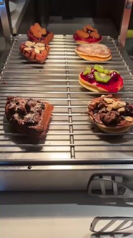 The video shows a close-up view of a metal cooling rack filled with various pastries. The pastries are arranged in rows on the rack, which appears to be inside a display case or oven. The pastries include heart-shaped waffles topped with chocolate and almonds, square pastries with fruit and jam, and round pastries with fruit and cream. The lighting is somewhat dim, creating a warm and inviting atmosphere. The camera slowly pans across the pastries, highlighting their textures and toppings.