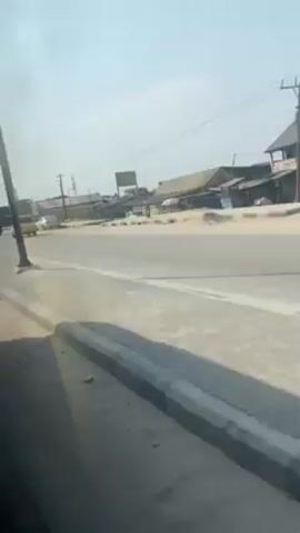 The video shows a view from inside a moving vehicle, looking out the side window at a street scene. The road is lined with buildings, some appearing residential and others commercial. There are utility poles along the roadside. Several vehicles pass by, including a motorcycle, a pickup truck, a van, and various cars. People can be seen walking along the side of the road. The weather appears to be clear and sunny. A sign is visible near one of the utility poles.
