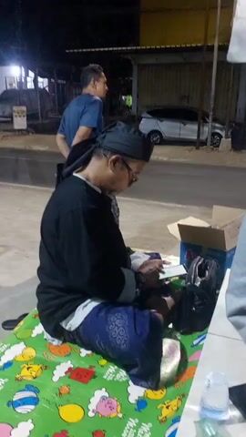 A man in a black shirt and black head wrap sits on a green mat decorated with cartoon characters. He is looking down at a book or electronic device in his hands. He is wearing blue patterned pants. Next to him is a cardboard box and a black bag. To his left, another man in a blue t-shirt stands and looks towards the left side of the frame. The scene appears to be outdoors at night, with some lighting from buildings in the background. A road with a vehicle is visible behind the men.