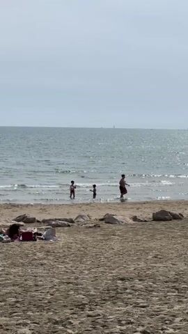 A beach scene shows three people, a man and two children, wading in the shallow water near the shore. One of the children is holding a soccer ball. Other people are lying on the sandy beach behind them, with beach gear scattered around. The water appears calm under an overcast sky. In the distance, a couple of sailboats can be seen on the horizon.
