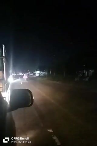 The video shows a nighttime view from inside a car driving on a road. The road is paved and illuminated by streetlights and the car's headlights. Other vehicles, including cars and motorcycles, are visible on the road ahead. The car's side mirror is visible in the foreground. The surroundings appear to be residential, with houses and trees lining the road. The video is labeled with the text "OPPO Reno8" and "2026.05.11 21:41" at the bottom of the frame. There is audio of people talking.