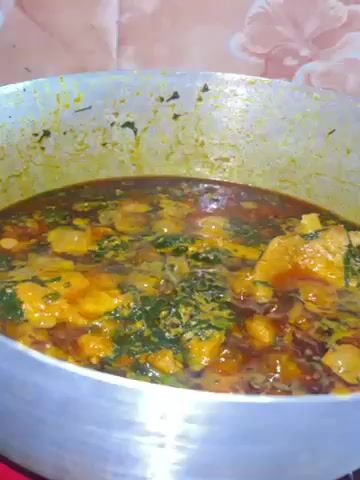 The video shows a close-up of a large, steaming pot filled with a thick, brown stew. Green leafy vegetables are mixed throughout, along with orange chunks of what appears to be meat or root vegetables. The pot itself is silver and shows signs of wear. The background features a tiled wall with a light brown and white pattern. The camera remains stationary throughout the brief video.