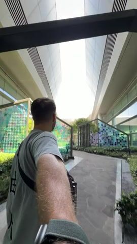 A man walks through a modern, brightly lit outdoor or semi-outdoor space in Jianyang City, China. The video is captured from a first-person perspective, with the man's right arm extended forward, holding the camera. He wears a light grey t-shirt and a dark cross-body bag, and is seen from behind. Behind him, a luggage trolley with a dark bag suggests he is traveling.

The architecture features sleek, modern buildings with white or light grey facades and prominent rectangular skylights that illuminate the pathway with strong, natural light, indicating a sunny day. The walkway is paved with hexagonal stone tiles. On either side of the path, decorative glass panels in shades of green, blue, and purple are arranged in a grid pattern, interspersed with green foliage and small trees in planters. The man is observing his surroundings as he walks, pushing his luggage. The sound of rolling wheels is audible, accompanying his movement through this contemporary setting, possibly an airport or a similar large-scale public facility. No discernible text is visible.