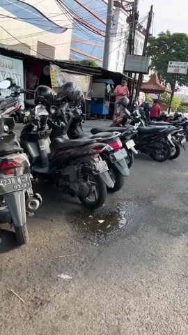 A man in a pink shirt and dark shorts stands on a sidewalk next to a row of parked motorcycles. Behind him, a building with a glass facade and an awning displays advertisements. The ground is paved with asphalt and concrete. The scene appears to be a street in a city, possibly in Southeast Asia given the style of the motorcycles and architecture.