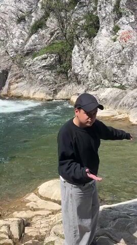 A person in a black sweatshirt, black cap and grey pants stands on a rocky outcrop by a pool of water, and waves their hand. The camera pans to show a small waterfall emptying into the pool, which is surrounded by grey rocky cliffs.