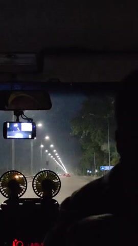 A first-person perspective video captures a nighttime drive on a road lined with streetlights. The road curves slightly to the right in the distance. Two small, circular fans with yellow blades are positioned on the dashboard in the foreground. A smartphone is mounted to the windshield, displaying a recording. The interior of the vehicle is dark, with the silhouette of a person visible in the passenger seat. Trees line the right side of the road, and traffic lights and signs are present. A red car is visible in the distance on the road ahead.