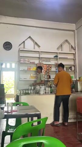 A person in a yellow t-shirt and black pants stands at a counter in what appears to be a small shop or eatery. They are facing a shelf filled with various items, including bottles, boxes, and what look like snacks. In front of the counter are stacks of plates and cups. To the left, another person in a green t-shirt is near a doorway, holding something. The environment is simply decorated with white walls and shelves. In the foreground, green chairs and a table are visible. The lighting suggests it's daytime.