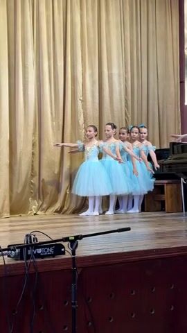 A group of young girls, dressed in identical light blue ballet costumes with golden embroidery and matching hair decorations, are performing on a wooden stage. The stage is set against a backdrop of golden curtains. The girls move across the stage, executing ballet steps and holding elegant poses with outstretched arms. Some girls are transitioning between movements while others stand still, maintaining their positions. In the foreground, a microphone on a stand with the brand name "VOTONE" is visible. The girls appear focused and graceful. The scene takes place indoors, suggesting a ballet recital or rehearsal in the urban settlement of Bilohirsk, Ukraine.