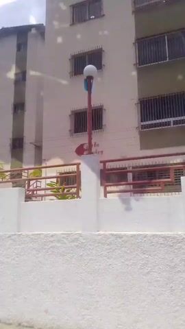The video shows a panning shot of an apartment building. The building is light-colored and has several windows, some with bars. There is a red railing in front of the building, and a sign that says "Castle Country". To the left of the building, there is a smaller building and a blue tarp. In the background, there are trees and a cloudy sky. The video also shows a wall and a person wearing a black cap. There is some writing on a white wall that appears to be a date and time.