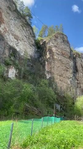 The camera slowly pans from a low angle upwards to capture a tall rock cliff formation on a sunny day with a few scattered clouds. The cliff is primarily tan with striations and some patches of green vegetation. There are some evergreen trees on the top of the cliff. At the base of the cliff is a grassy area separated by a green mesh fence. Some people are gathered near the base of the cliff behind the fence. The camera continues to pan to the right, showing a dirt path running along the edge of a body of water. A group of people are walking along the path, and a forest is visible on the far side of the water. A zip line is visible high above the cliff.