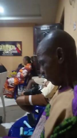 The video shows the interior of a room where a group of people are sitting, presumably at an event or gathering. The camera focuses initially on an older man with a bald head, wearing a patterned purple and green shirt with a tan panel in the front. He is sitting on a blue plastic chair, looking down. Other people can be seen seated in the background, though they are somewhat blurry. The room has painted walls and appears to have a simple decor. Musical sounds can be heard in the background. The camera then pans to show a wider view of the room, capturing more of the seated audience and the white plastic chairs they are seated on.