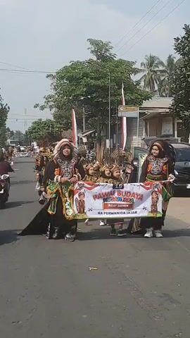 A group of people are walking in a parade. The people in the front are wearing elaborate traditional costumes with headdresses. They are holding a banner that says "PAWAI BUDAYA NUSANTARA ADAT DAYAK RA PERWANIDA JAJAR". Behind them are children also in traditional attire. They are walking down a paved road with trees and buildings on either side. There are also motorcycles and cars visible in the background. The sky is clear and the sun is shining.