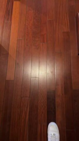 This media file shows a walkthrough of a hotel room. The room has polished wooden floors throughout. The first shot is a close-up of the floor, with a white shoe visible at the bottom. The camera then pans to reveal a hallway leading into the main room. In the hallway, there is a dark suitcase with a black backpack on top, and a small trash can. Further into the room, a dining area with a table and chairs is visible, as well as a large window with striped curtains. The camera continues to move, showing a television on a wooden stand, a lamp, and a portion of a bed. In the background, there are more chairs and windows. The overall impression is of a well-appointed hotel suite. There is no visible text in the media. The lighting suggests it is daytime, possibly late afternoon, given the warm tones of the interior. The setting appears to be a hotel room in La Gaspara, Spain, though no explicit confirmation of this location is given within the video itself.