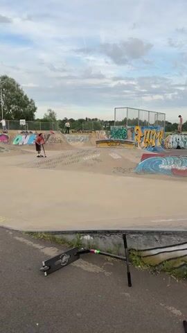 The video takes place in a skateboard park on what appears to be a partly cloudy day. The park is filled with various ramps, platforms, and graffiti art. Several people are present, some observing and others actively using the park. A young man, shirtless and wearing shorts and Adidas sneakers, is riding a scooter with pink handlebars. He navigates through the park, performing a stunt on a small quarter pipe. The fallen scooter is lying on the ground.