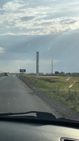 The video shows a roadside view from inside a vehicle. The road stretches ahead, with a grassy field to the right. A tall, white sign with vertical red lettering is visible on the right side of the road. The letters appear to spell out "TIMIRIAZEVO". In the distance, there is a billboard with blue and white lettering, and a few scattered buildings and utility poles. The sky is partly cloudy, with sunlight streaming through. A few small particles are visible on the windshield.