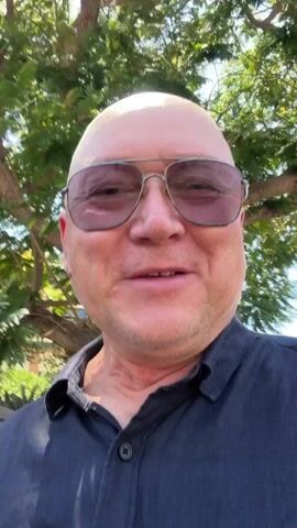 A medium shot captures a bald man wearing aviator sunglasses and a dark collared shirt. He is smiling directly at the camera, with lush green trees and dappled sunlight forming the background. The man's head is tilted slightly forward, and his expression is animated as he speaks. The camera appears to be held close to him, giving the impression of a selfie or a personal video message.