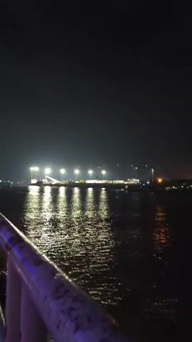 A nighttime video captures a harbor or industrial waterfront. Bright spotlights illuminate an area on the shore, where cranes and shipping containers are visible. The water in the foreground reflects the lights, creating a shimmering effect. Several small boats or vessels are present on the water, some appearing to be moving. The overall scene suggests an active port or industrial site operating after dark.