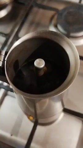 A close-up shot shows a metal stovetop espresso maker on a gas stove. The top chamber of the espresso maker is filled with dark liquid, presumably coffee, and small bubbles are forming on the surface, indicating it is heating. The espresso maker is silver and has a handle on the left side. The gas stove grates are visible, as are the burners, one of which is partially visible in the background. The lighting is dim, suggesting the shot was taken indoors, likely in a kitchen.