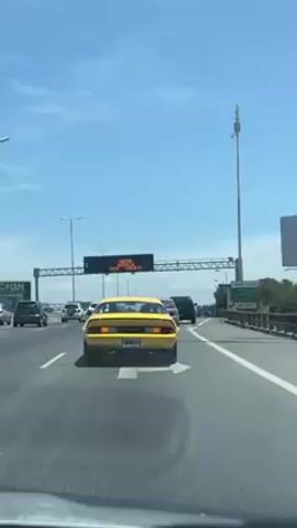 The video shows the rear view of a yellow Chevrolet Camaro driving on a highway with several lanes.  There are other vehicles on the road, and an overhead electronic traffic sign displaying information. The sky is blue with some clouds. There is road infrastructure visible, including lampposts, towers, and highway signs. The camera is inside a car.