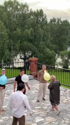 The video shows a group of five adults, three men and two women, on an outdoor patio area. They are engaged in a game where they are blowing up balloons. Two men are holding blue and white balloons respectively, with their mouths on them as if inflating them. A woman in a black and white polka dot dress is actively blowing into a white balloon, her cheeks puffed out. Another man, wearing a light-colored shirt and pants, stands nearby, appearing to watch the activity. A fifth man, dressed in a dark suit, is moving away from the group, his mouth open as if he is about to pop a balloon or has just done so. The patio is paved with decorative stone tiles and is bordered by a black metal railing. Beyond the railing, there is a lush green landscape with tall trees and a body of water, possibly a lake or river, in the background. The sky appears overcast. Two individuals in the foreground, partially visible, are holding cameras and appear to be recording the event.