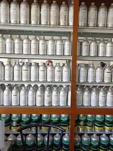 The video pans across shelves filled with rows of metallic bottles, likely containing liquids, such as perfumes or chemicals. The bottles are of various sizes and are arranged neatly on glass shelves within wooden display units. In the background, a person is partially visible, seemingly interacting with items on a counter. The lighting suggests an indoor environment, possibly a shop or storage area. The overall impression is one of organized retail or inventory.