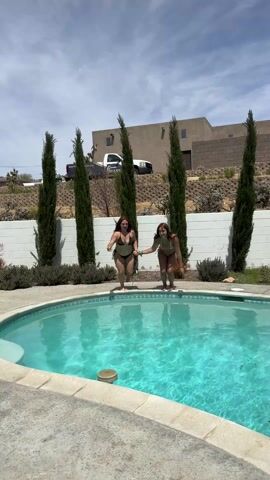 The scene depicts two young women in bikinis jumping into a backyard swimming pool on a sunny day. They leap simultaneously from the pool's edge, creating a large splash upon impact. Both react with surprise and exclamation after submerging. The pool is surrounded by a concrete deck, lush greenery, and tall, slender trees. In the background, there is a stucco house and a truck parked along a hillside.