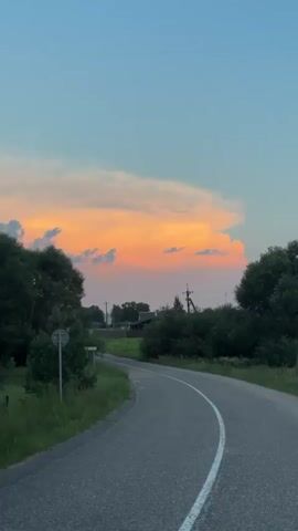This is a vertical video clip filmed from a vehicle traveling along a winding asphalt road in a rural setting. The road is bordered by lush green grass and trees on both sides. In the distance, a small village with houses and power lines can be seen. The sky above is a gradient of blue and orange, with fluffy clouds illuminated by the setting sun.

The time of day is clearly sunset, indicated by the vibrant colors of the sky and the soft lighting. The weather appears clear and pleasant, with no visible precipitation. The location is identified as Нёманскі сельскі Савет, Belarus.

There is a white road sign on the left side of the road with text that reads "МАЛОЧКА" in white letters on a dark background. This sign likely indicates the name of a locality or a direction. The overall mood of the video is serene and peaceful, capturing the beauty of a rural landscape at dusk. No people or specific activities are visible, focusing solely on the natural scenery and the journey.