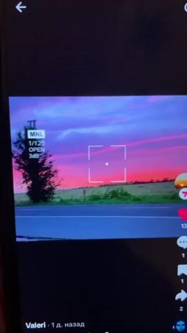 The video shows a digital display on a mobile device. A scenic video is playing on the screen, featuring a sunset over a rural landscape. The sky is a vibrant mix of pink, purple, and blue, with streaks of pink clouds. In the foreground, there is a road, a field of green grass, and a lone tree on the left side of the frame. A circular overlay with a number 7 is visible on the right side of the screen, as is a heart icon with the number 13. The interface elements of a social media application, likely TikTok, are present, including a back arrow, a search icon, and icons for likes, comments, shares, and a profile. The video appears to be recorded from another device, as evidenced by reflections and the edge of the recording device's screen. The bottom of the screen displays text indicating a username "Valeri" and a timestamp "1 д. назад" (1 day ago in Russian).