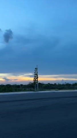 This is a short video capturing a scenic view of the sky and landscape at dusk in Mui Ne, Vietnam.

The scene is dominated by a vast sky, a gradient of deep blue at the top transitioning to lighter blues and hints of orange and yellow near the horizon. The horizon itself is marked by a line of dense green foliage, suggesting a natural landscape or a cultivated area. In the foreground, a dark asphalt road with white markings is visible, indicating a man-made structure like a street or parking lot.

Scattered throughout the landscape are a few slender trees, some appearing young and sparse, others more established. Prominently featured are two streetlights, tall and thin with single fixtures at the top. One streetlight is in the middle distance, and another is further to the right. A small, red fire hydrant is also visible near the right edge of the frame.

The time of day is clearly dusk or twilight, with the soft, diffused light and the colors in the sky strongly indicating this. The clouds are a mix of dark, moody masses and thinner, wispy formations. The overall mood is one of peacefulness and natural beauty, with the vastness of the sky and the quiet landscape creating a serene atmosphere. There are no people visible in the video. No text is visible.