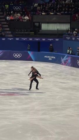 In a wide shot, two figures ice skate on a rink, engaged in pairs figure skating. The arena seating is largely full of an audience, all facing toward the ice rink. Behind the rink is blue and purple signage for the 2026 Milano Cortina Olympic games. The male figure skater wears all black; his partner wears brown and black. She is raised in the air, parallel to the ice. They hold each other. The ice has some light scratch and scuff marks and some pink circular designs. Two figures stand in the background, looking at the figure skaters.