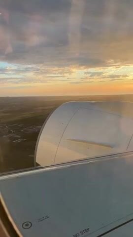 This video shows a view from an airplane window during sunset. The wing of the airplane is visible in the foreground, with the engine casing to the right. The sky is filled with dramatic clouds, with the sun casting a warm orange glow on the horizon. The landscape below is a blur of green fields and scattered buildings, suggesting the plane is either taking off or landing. The camera is stable, indicating it is likely mounted or held by a passenger.