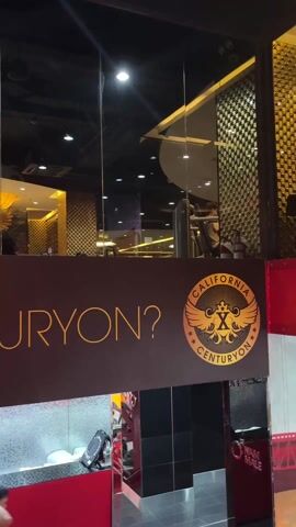 Here's a detailed description of the video:

The video shows the exterior of a gym called "California Centuryon." The camera is positioned at street level, looking up towards the gym's entrance, which has a large glass window. The gym's signage is visible, with the name "California Centuryon" and a logo featuring wings and a stylized "X". The signage is partially obscured.

Inside the gym, various workout machines and people exercising can be seen through the glass window. The interior design features a modern aesthetic with gold and black accents.

A person wearing a white shirt is briefly visible near the bottom left corner of the frame, their head down. The overall environment appears to be a busy urban area. The lighting is bright, likely from both natural light and interior lighting within the gym. There is a red structural element in the foreground, adding a pop of color to the scene.