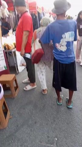 The video depicts a busy outdoor market. In the foreground, several people are walking. A man in a red shirt and hat walks left, holding plastic bags. To his right, a woman in a patterned outfit and a man in a blue t-shirt and shorts walk away from the camera. Further back, a young girl with long black hair is visible. The market stalls are lined with clothing and other goods. The ground is asphalt. The overall atmosphere appears to be crowded and active.