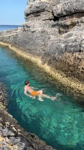 A young man is swimming in a narrow, turquoise inlet bordered by steep, rocky cliffs. He is wearing orange swimming trunks. A young woman joins him, swimming in the same inlet. The water is clear, revealing the rocky bottom. The sky is blue and the sun is shining.