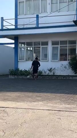 A person is sweeping in front of a white building with blue trim and multiple windows. The person is wearing a black shirt and shorts and is holding a broom. The ground in front of the building is paved with interlocking bricks. There is some greenery near the building.