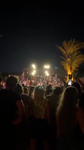 A nighttime outdoor scene shows a large crowd of people gathered in front of a stage decorated with lights and geometric patterns. Palm trees are visible on the right side of the frame. The sky is dark. People in the foreground are silhouetted. The crowd appears to be enjoying an event, possibly a concert or festival.