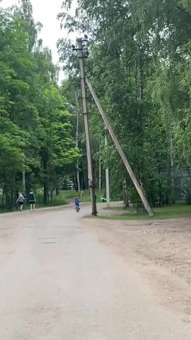 The video shows a dirt road lined with trees. On the left side of the road, two people walk away from the camera. Further down the road, two children on bicycles ride towards the camera. A utility pole leans precariously at an angle to the right of the road. The sky is overcast.