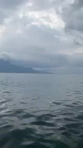 The video shows a body of water, likely a lake or sea, under a cloudy sky. The water is slightly choppy with gentle ripples. In the distance, land with a mountain range is visible, shrouded in a layer of mist or cloud cover. The overall scene suggests a cool, overcast day.