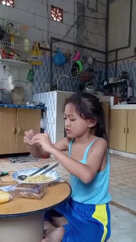 A young girl is sitting on the floor at a small round table in a kitchen. She is wearing a light blue tank top and blue shorts with a yellow stripe. On the table in front of her are a plate of noodles, a wrapped cob of corn, and a package of what appears to be dessert. She is holding her phone and looks up and to the right before looking back down at the phone. Behind her are kitchen cabinets and a tiled wall. A red door is visible in the background.