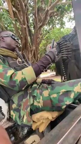 A medium shot captures a soldier clad in camouflage uniform and dark sunglasses. He is seated beside a heavy machine gun mounted on an armored vehicle or structure. The soldier's gloved hands are resting near the gun’s mechanism. A tree is visible in the background.
