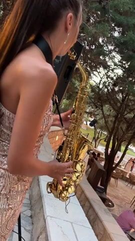 A young woman with long, dark brown hair pulled into a high ponytail, wearing a sparkly, rose-gold sequined dress, plays a saxophone. She is positioned outdoors on a stone ledge, with a speaker stand to her left. Behind her, trees with dense foliage are visible, along with glimpses of an outdoor dining area with tables and chairs, suggesting a restaurant or event venue. People are seen in the background, some seated, others standing, engaged in conversation or attending the event. The lighting suggests it is late afternoon or early evening. The woman appears focused and engaged in her performance. A speaker with the visible text "JBL" is to the left of the woman. The setting, with its outdoor dining and natural surroundings, combined with the formal attire of the performer, hints at a special occasion or a live music performance at a scenic location.