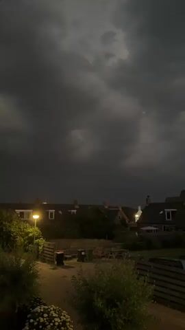 The video shows a view of houses at dusk with dark, cloudy skies overhead. A streetlamp on the left illuminates a pathway between two houses. In the foreground, there are bushes and some white flowers. The sky appears stormy, with heavy, dark clouds moving across it. There are no people visible in the video. The overall atmosphere is dark and moody, suggesting the approach of a storm.